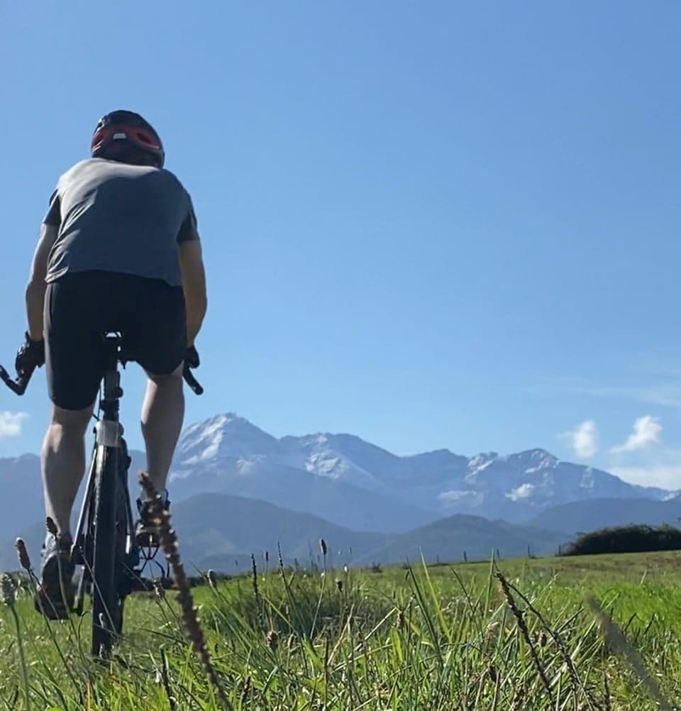 Seen from behind, a rider pedals across a grassy field toward snow-dusted peaks.